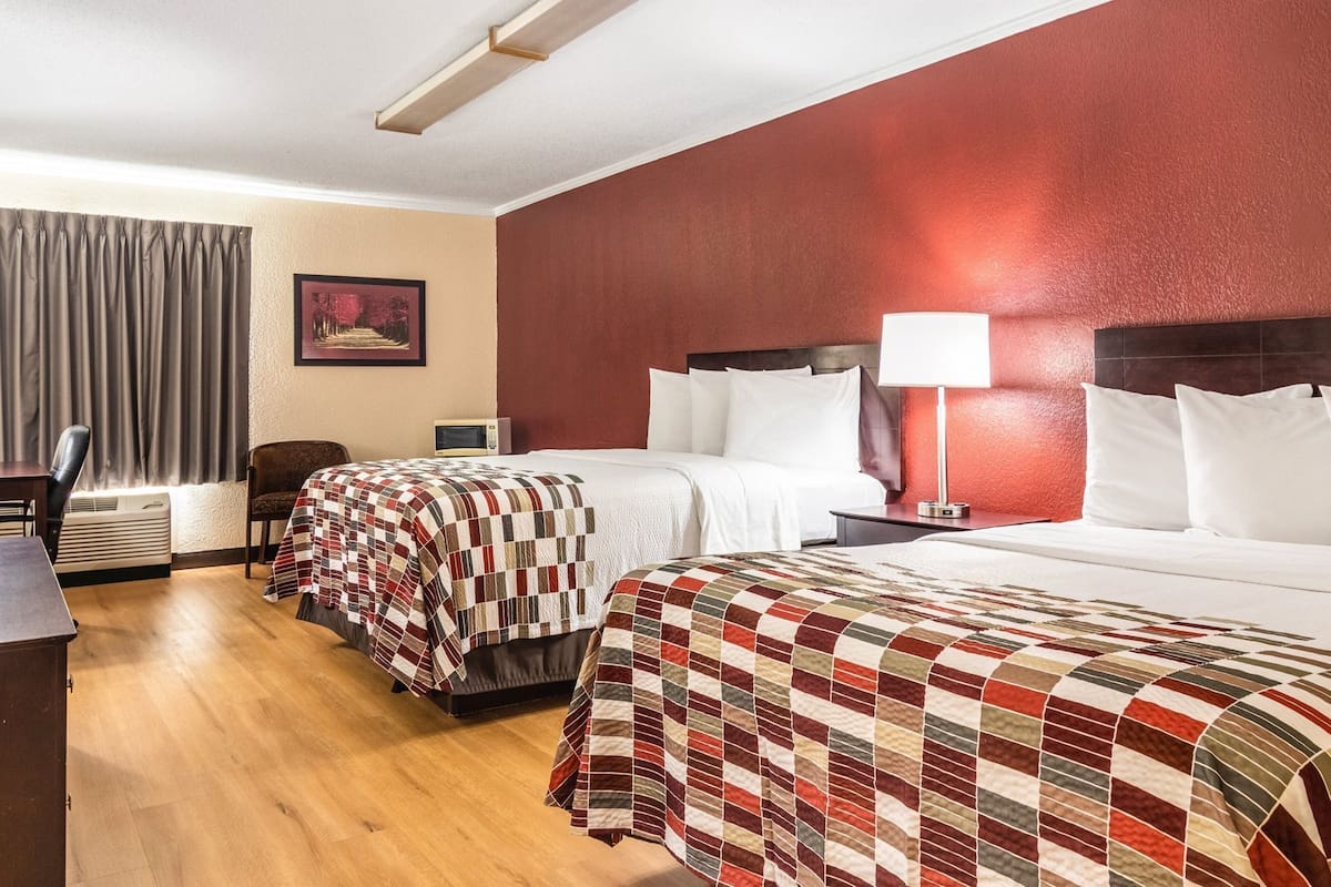 A hotel room with two beds, a desk, a chair, an air conditioning unit, and a lamp.