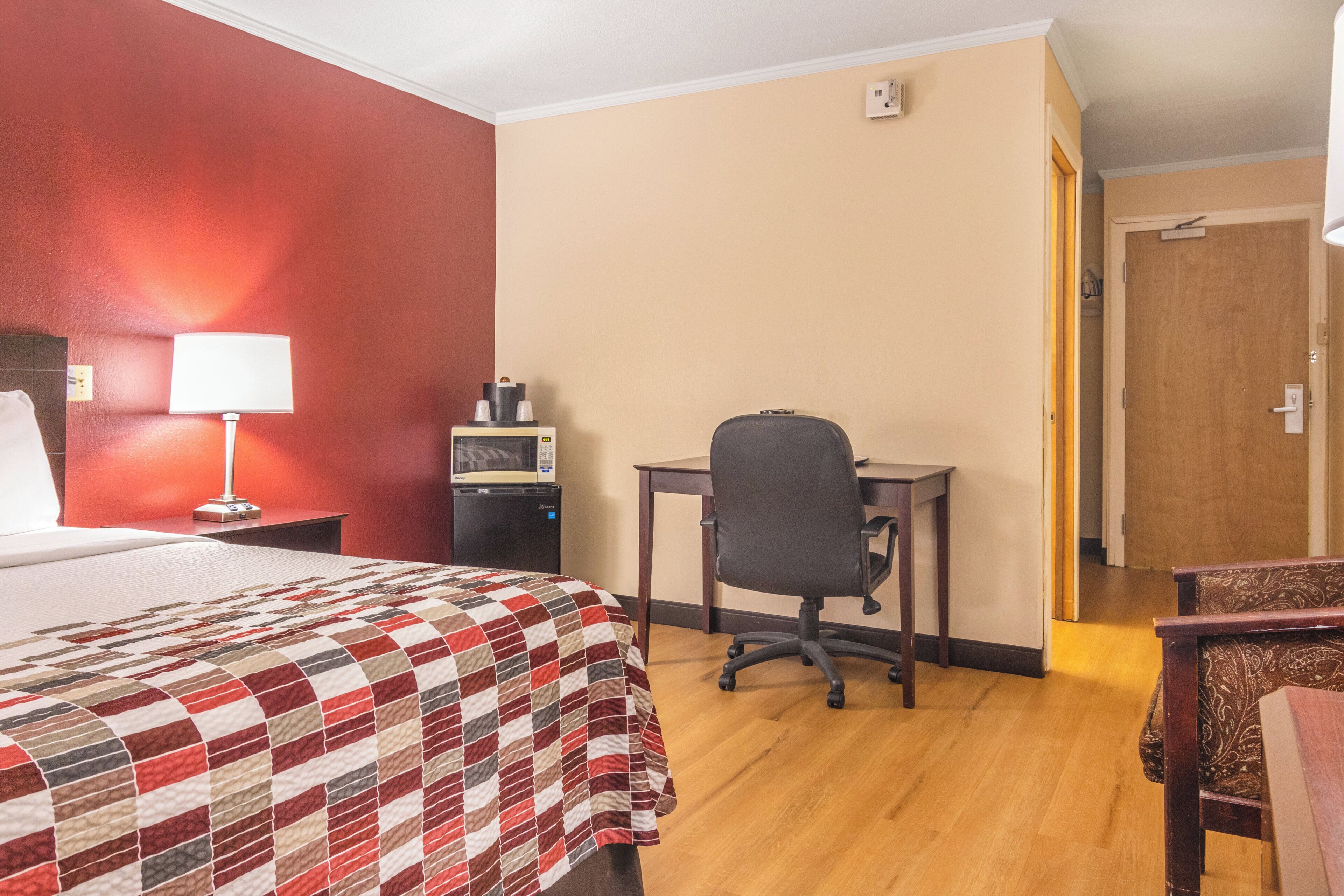 Quarto de hotel com cama, escrivaninha, cadeira, luminária e micro-ondas.