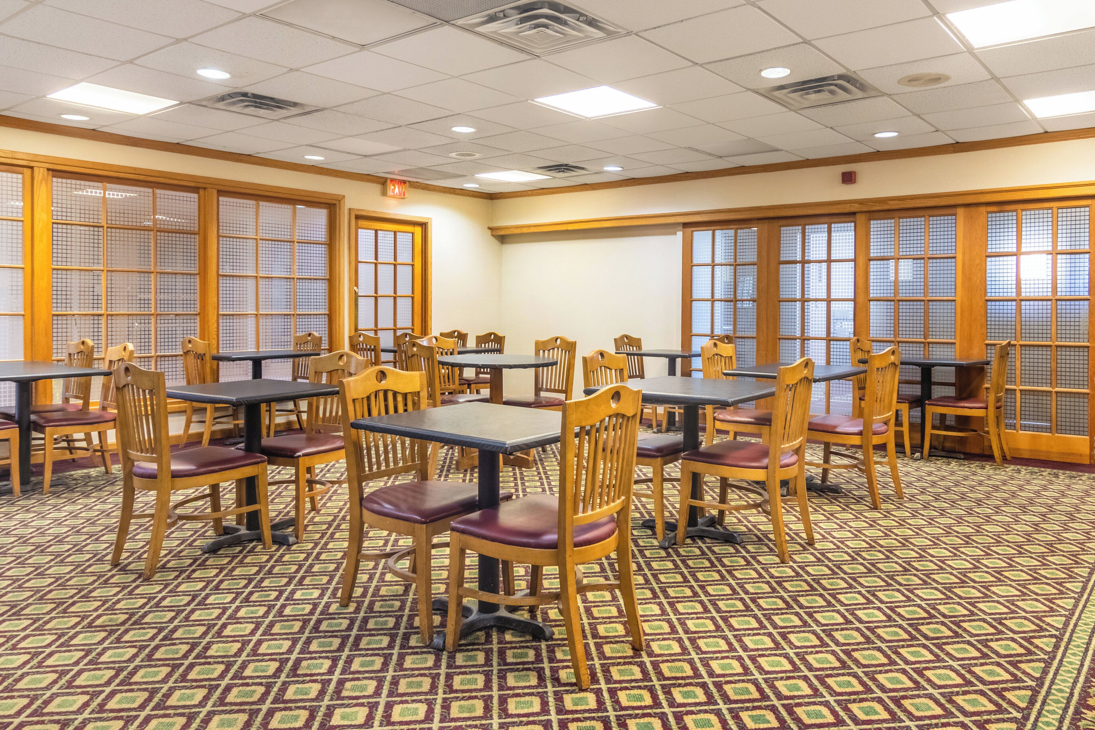A dining area with multiple tables and chairs, patterned carpet, and large windows with wooden frames.