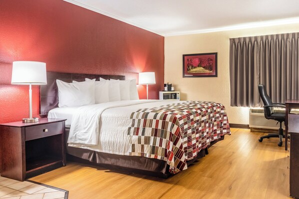 A hotel room with a bed, a desk, a chair, a nightstand, a lamp, a picture on the wall, and a window with curtains.