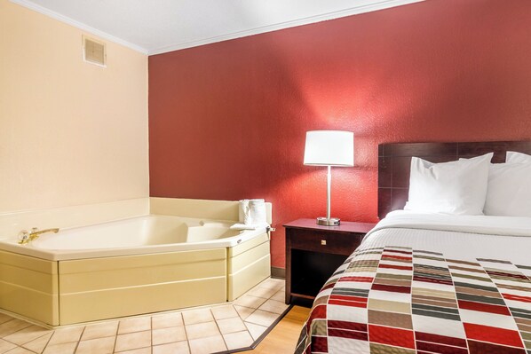 Habitación de hotel con bañera, cama con almohadas, mesita de noche con lámpara y pared roja.