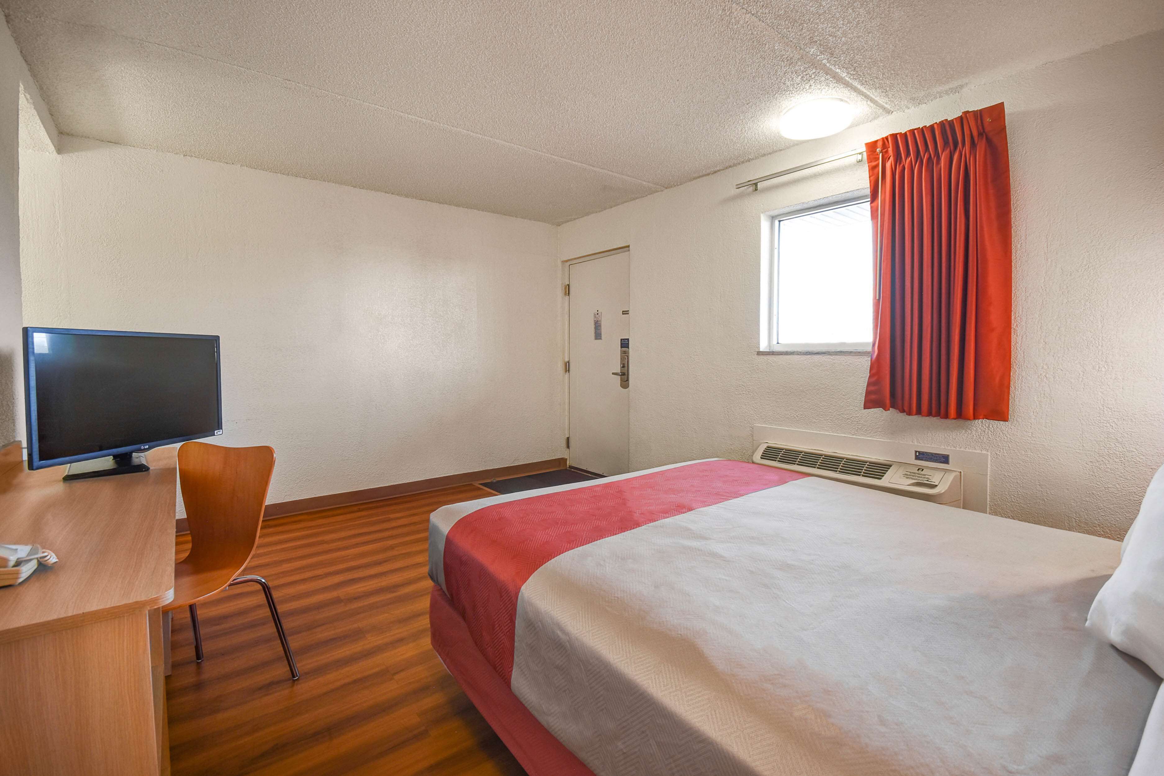 Habitación de hotel con cama, escritorio con computadora, silla, ventana con cortinas y unidad de aire acondicionado.
