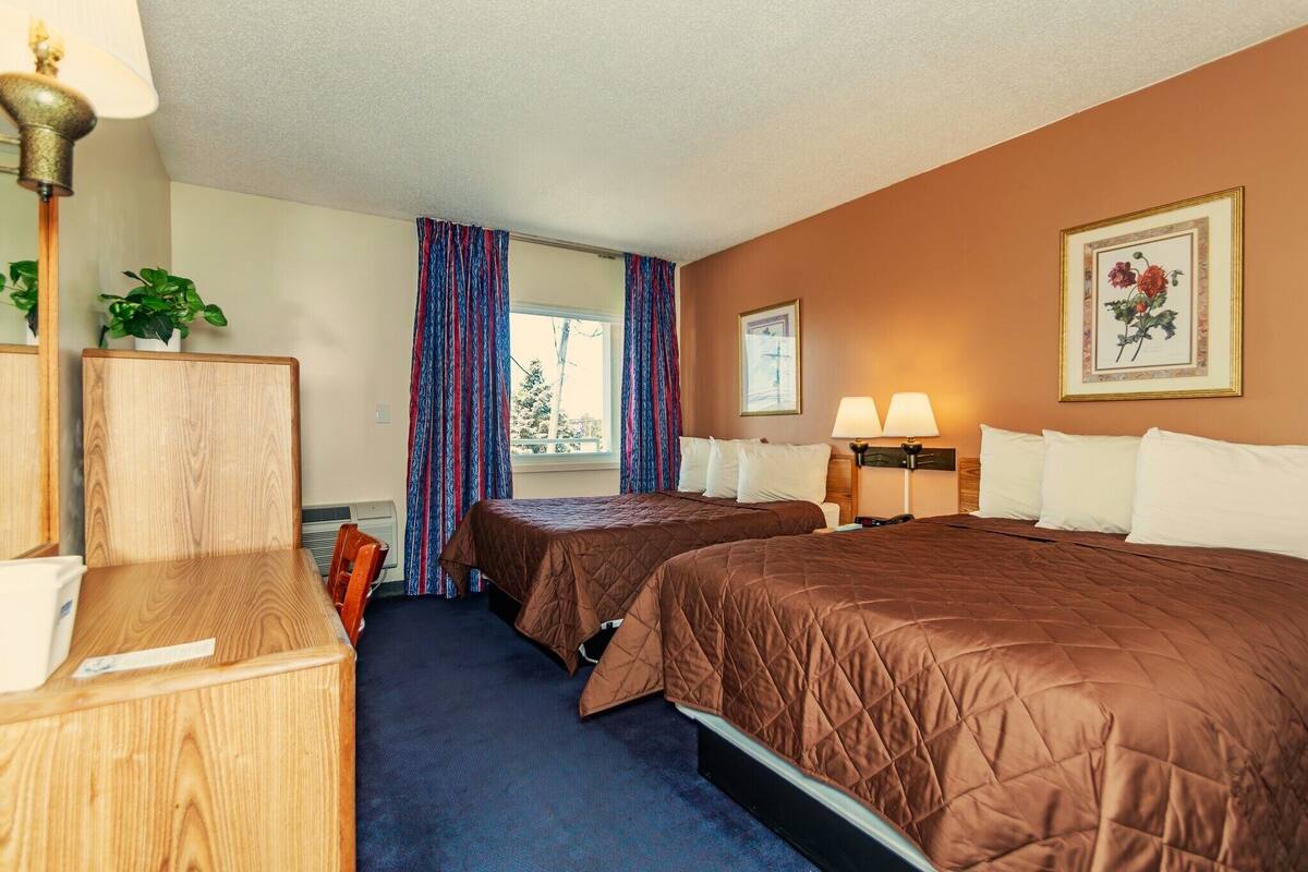 A hotel room with two beds, a desk, a lamp, a plant, and a window with curtains.