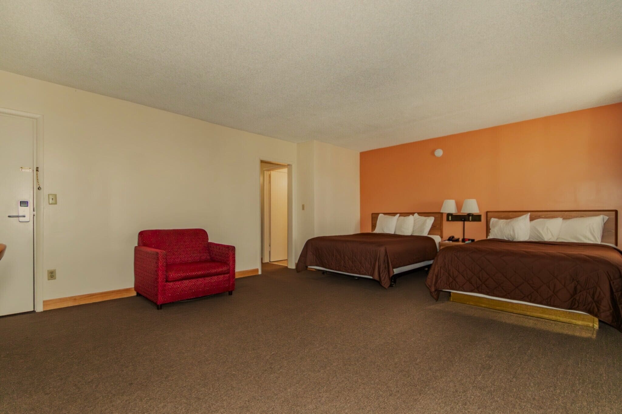 A hotel room with two beds, a red armchair, and a door.