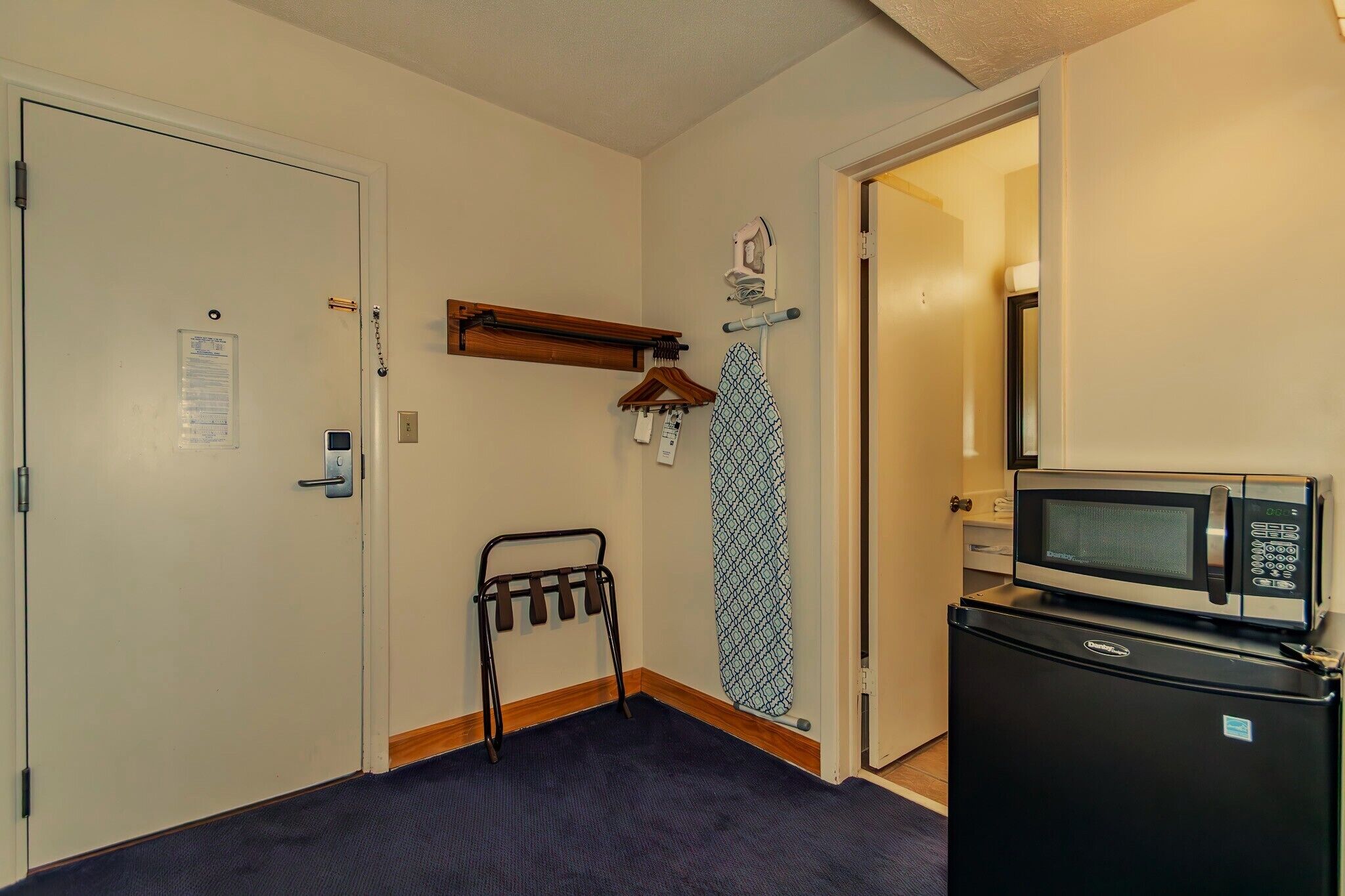 A compact room with a bed, a small refrigerator, a microwave, and a door with a coat rack and ironing board.
