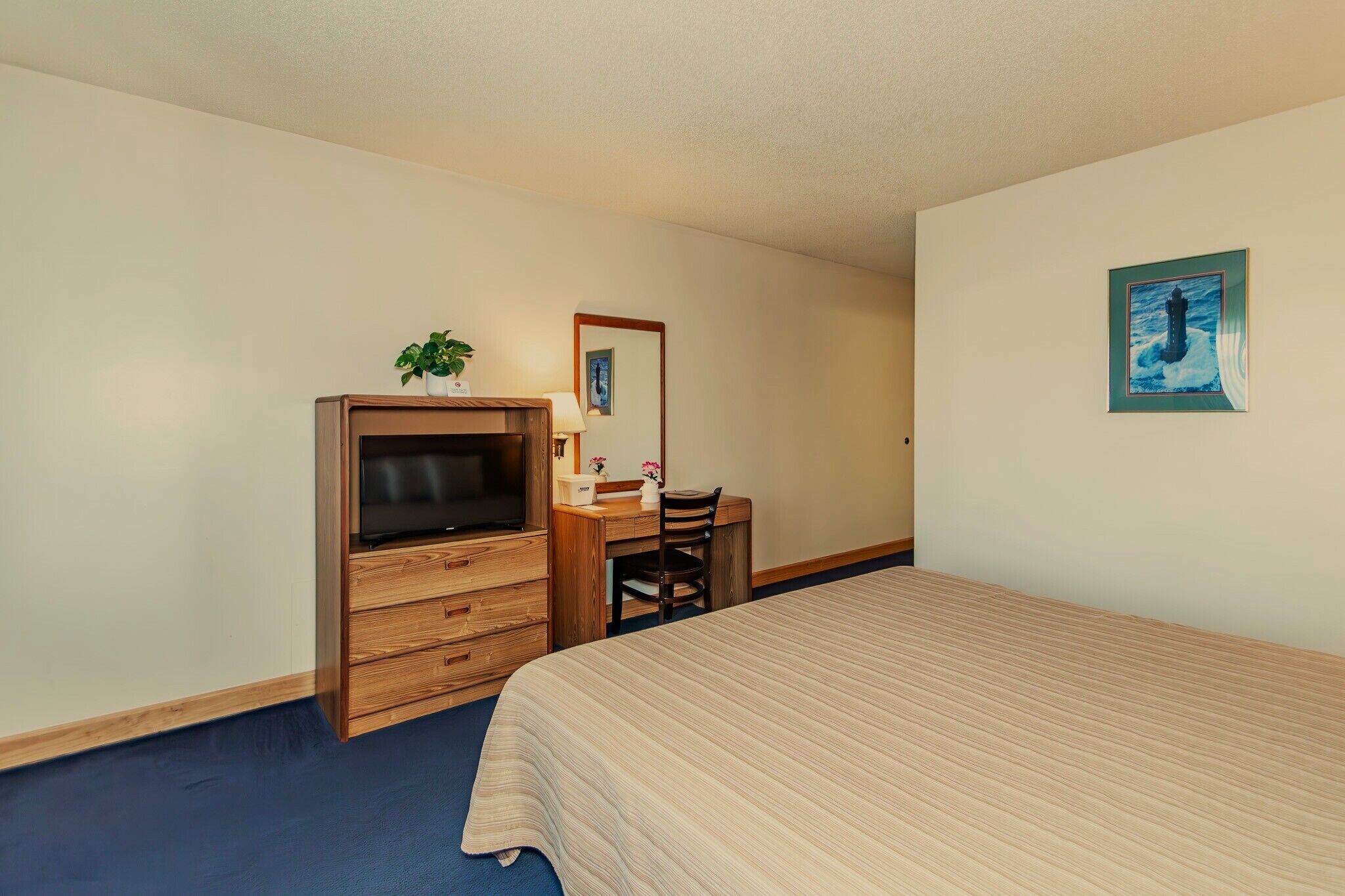 A bedroom with a bed, a wooden dresser with a television, a desk with a chair, and a wall-mounted painting.