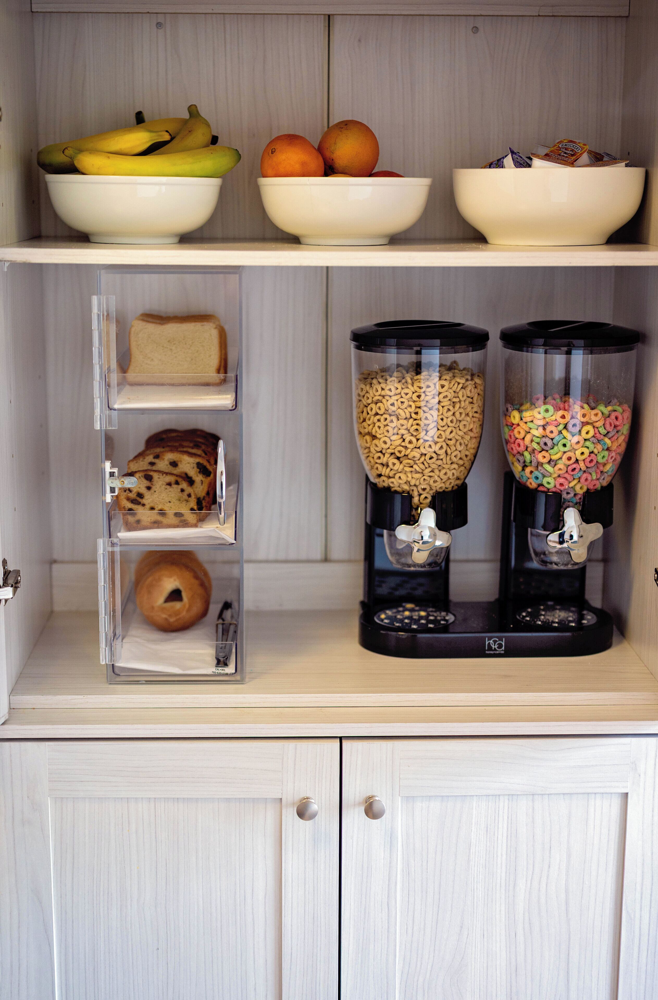 A kitchen with a bread shelf, a fruit bowl, and two cereal dispensers.