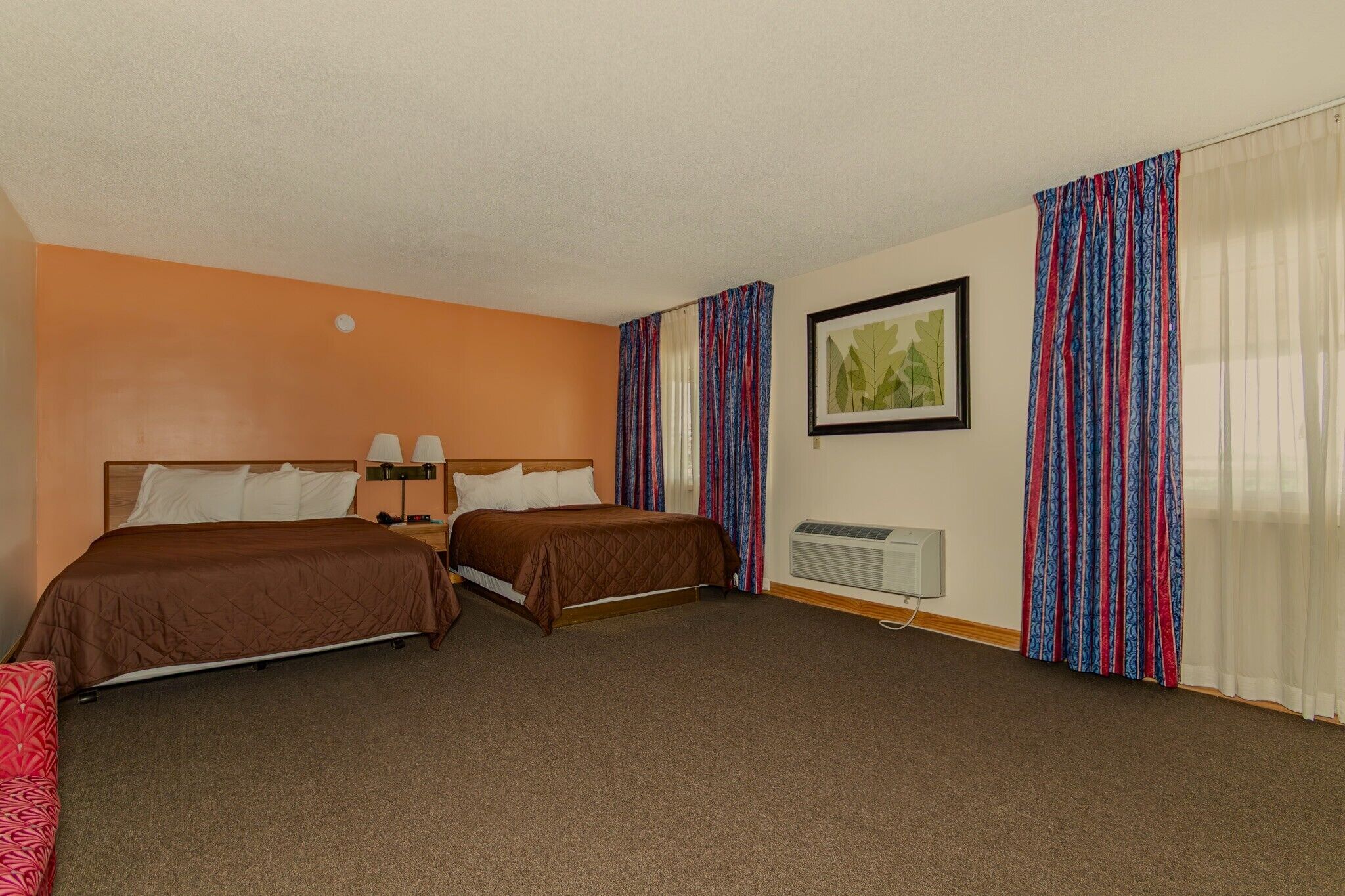Quarto de hotel com duas camas de solteiro, uma mesa de cabeceira, um abajur, um quadro emoldurado e uma janela com cortinas estampadas.