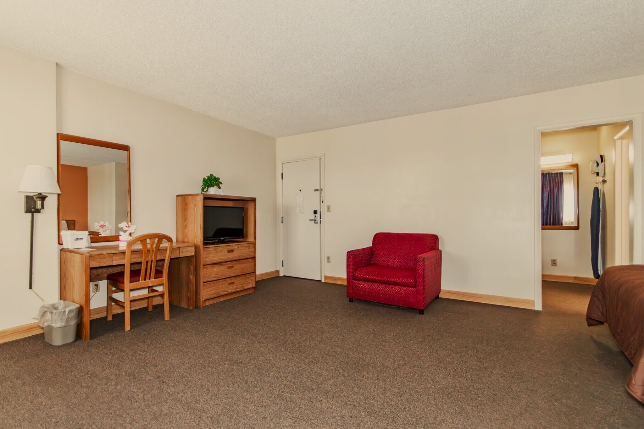 A compact hotel room with a wooden desk, a red sofa, a television on a wooden cabinet, and a door leading to another room.