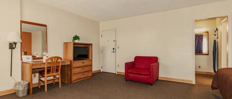 A compact hotel room with a wooden desk, a red sofa, a television on a wooden cabinet, and a door leading to another room.