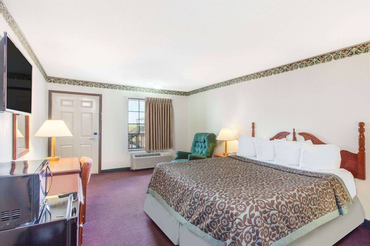 Quarto de hotel com cama, escrivaninha, cadeira, televisão e janela com cortinas.