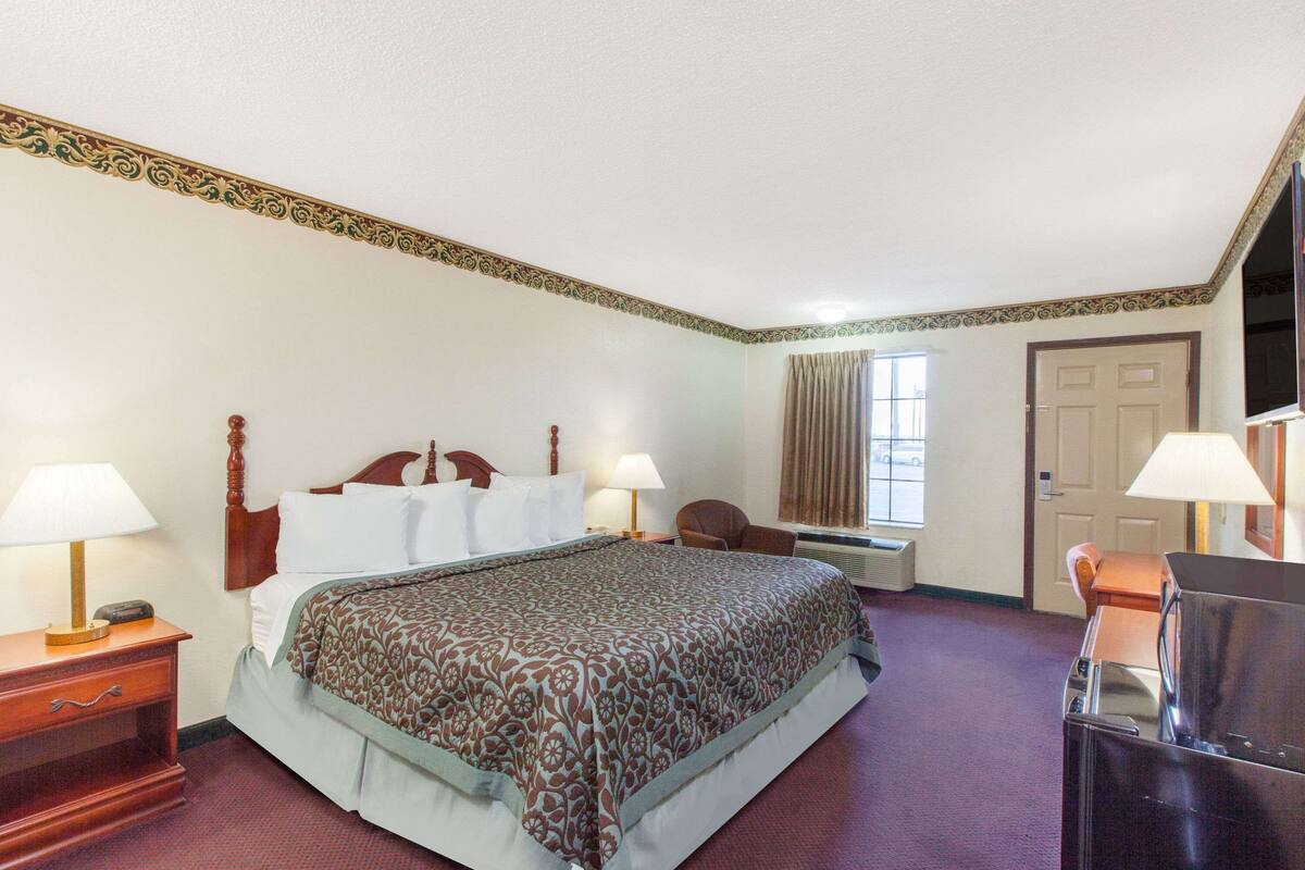 Quarto de hotel com uma cama grande, duas mesas de cabeceira com luminárias, uma televisão, uma escrivaninha com cadeira e uma janela com cortinas.