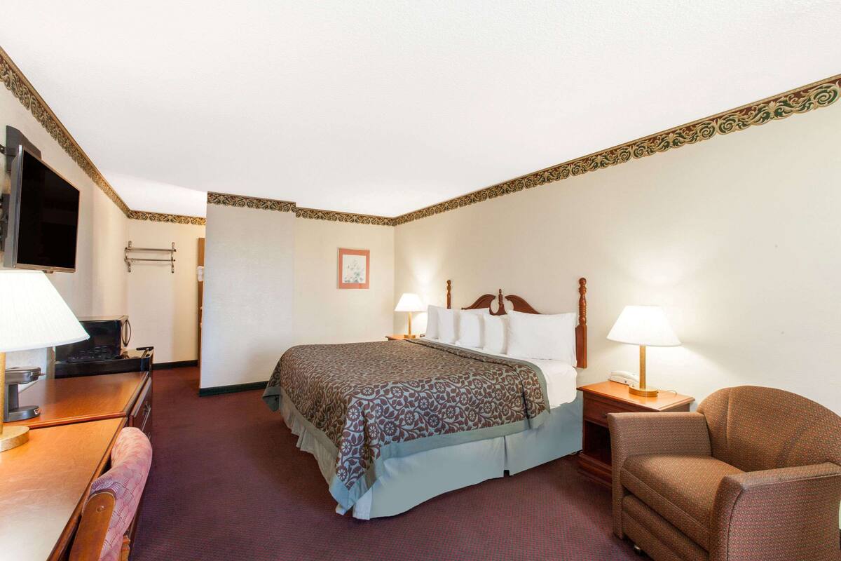 Quarto de hotel com cama, duas luminárias de cabeceira, uma cadeira, escrivaninha com luminária, televisão e um quadro na parede.