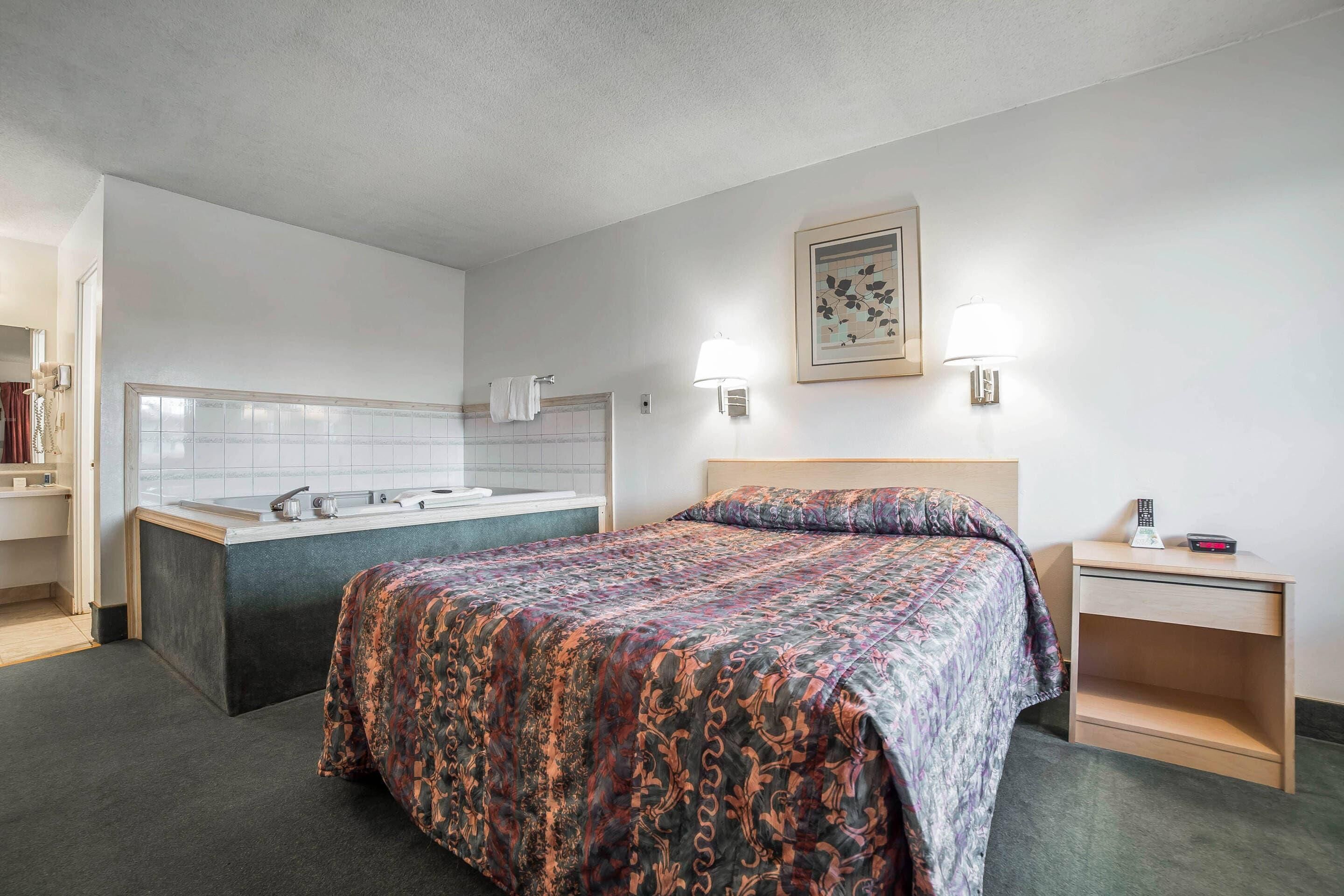 A hotel room with a bed, a small bedside table, a nightstand, a wall-mounted lamp, a framed picture, and a bathroom with a bathtub and sink.