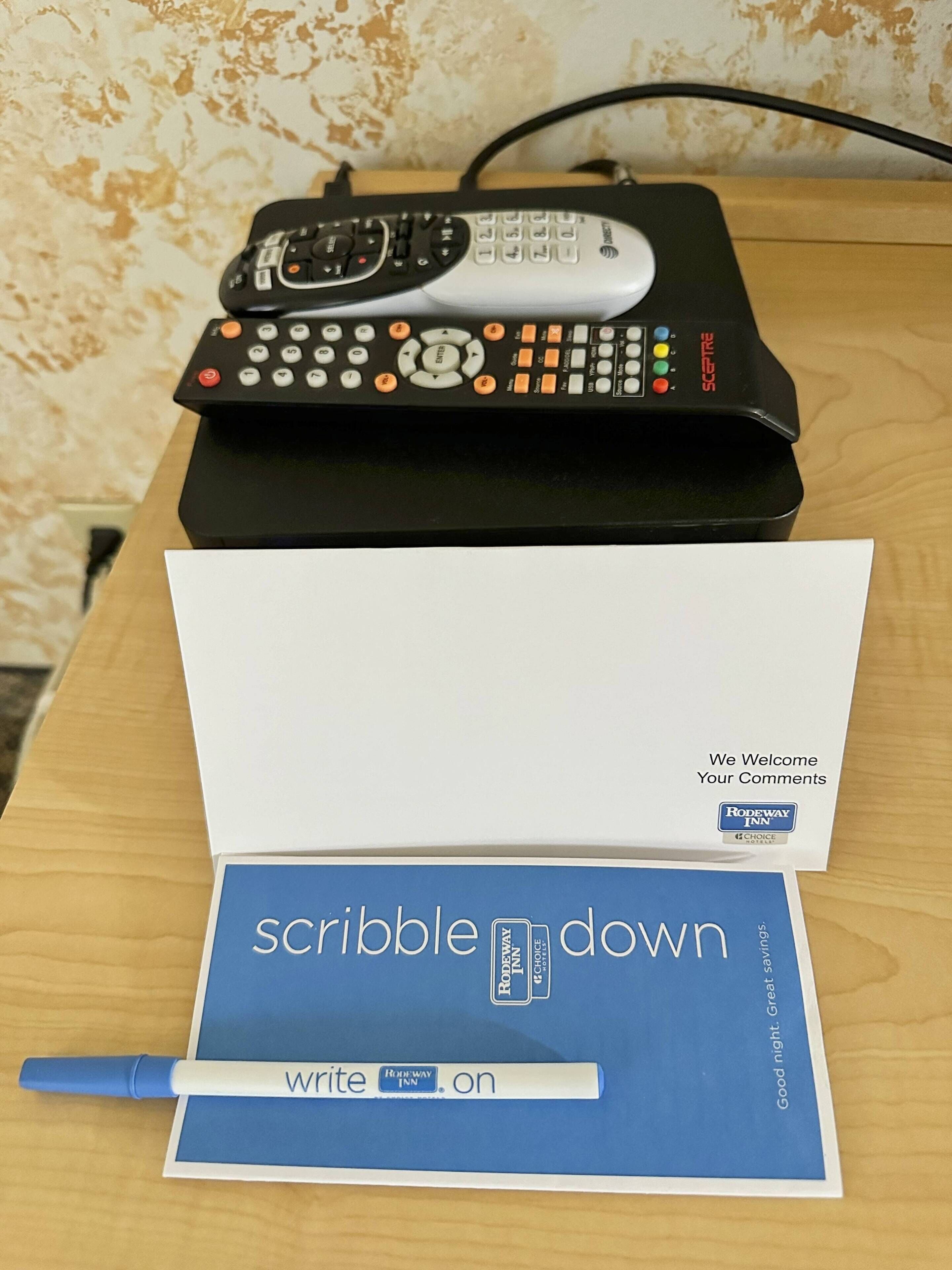 Eine Fernbedienung, eine weiße Karte mit der Aufschrift "We Welcome Your Comments" und "RODEWAY INN" sowie eine blaue Karte mit "scribble down" und "write on".