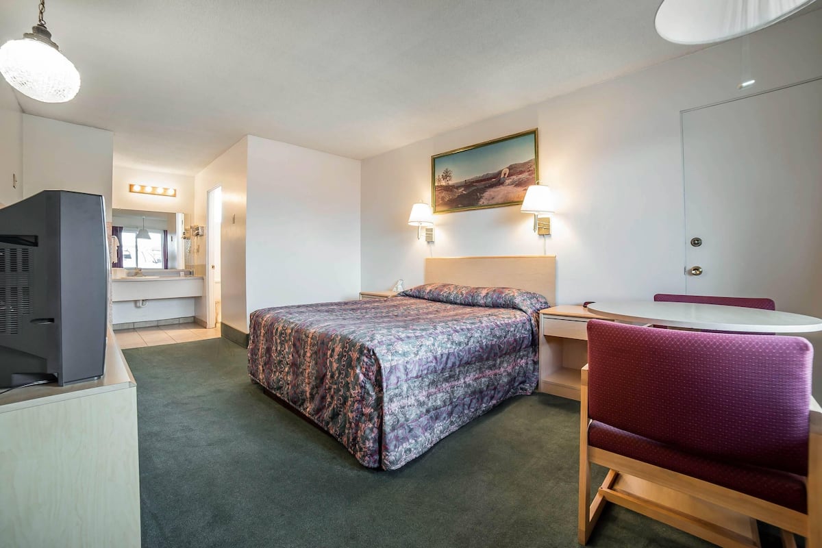 A hotel room with a bed, a desk, a chair, a television, a painting on the wall, and a door.