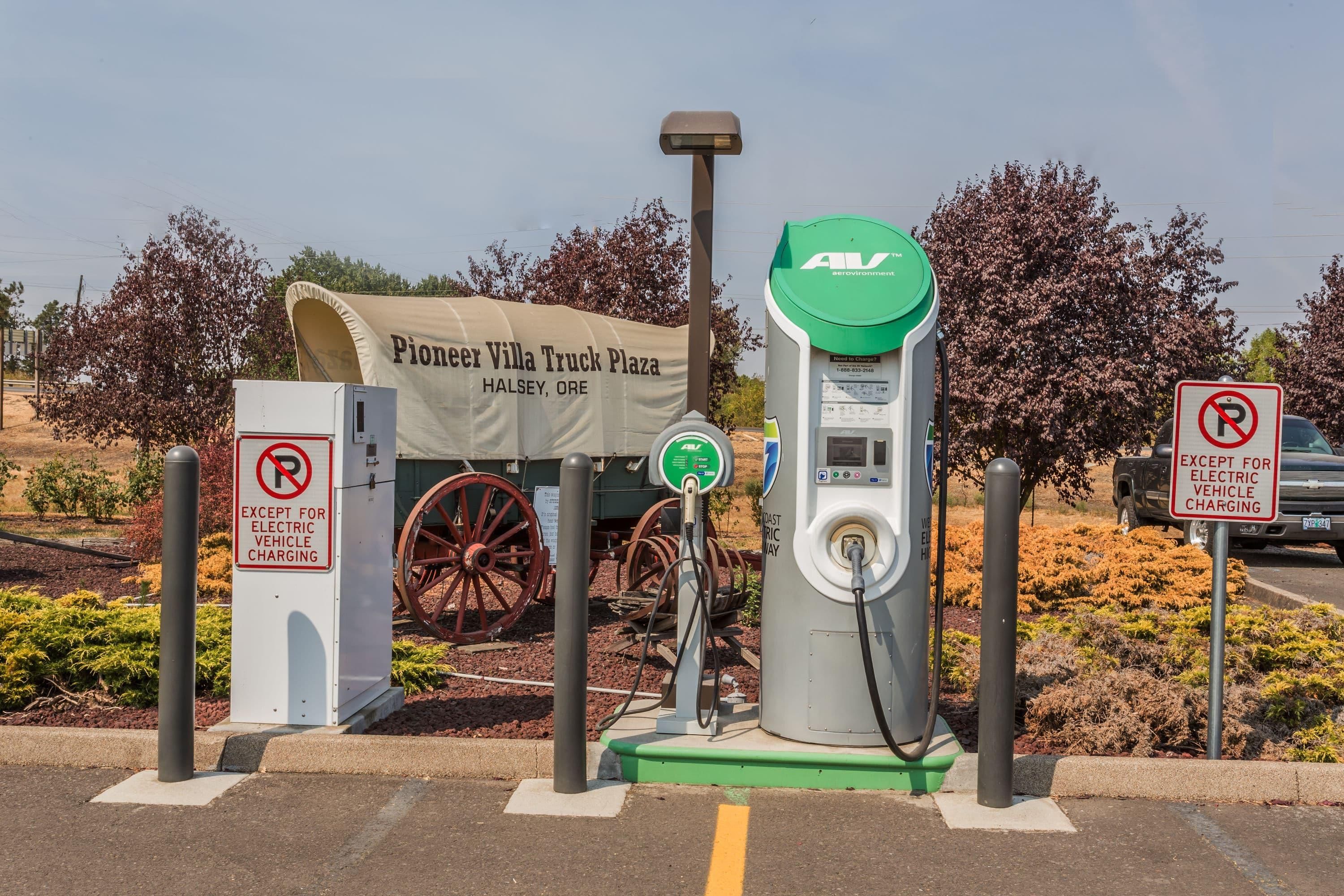 Una estación de carga para vehículos eléctricos con un cartel que dice 'Pioneer Villa Truck Plaza Halsey, Ore' y un letrero de 'prohibido estacionar' excepto para vehículos eléctricos en carga.