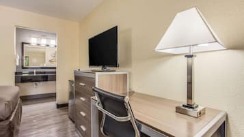 A hotel room with a wooden desk, a black chair, a flat-screen TV, a lamp, a mirror, and a kitchenette.