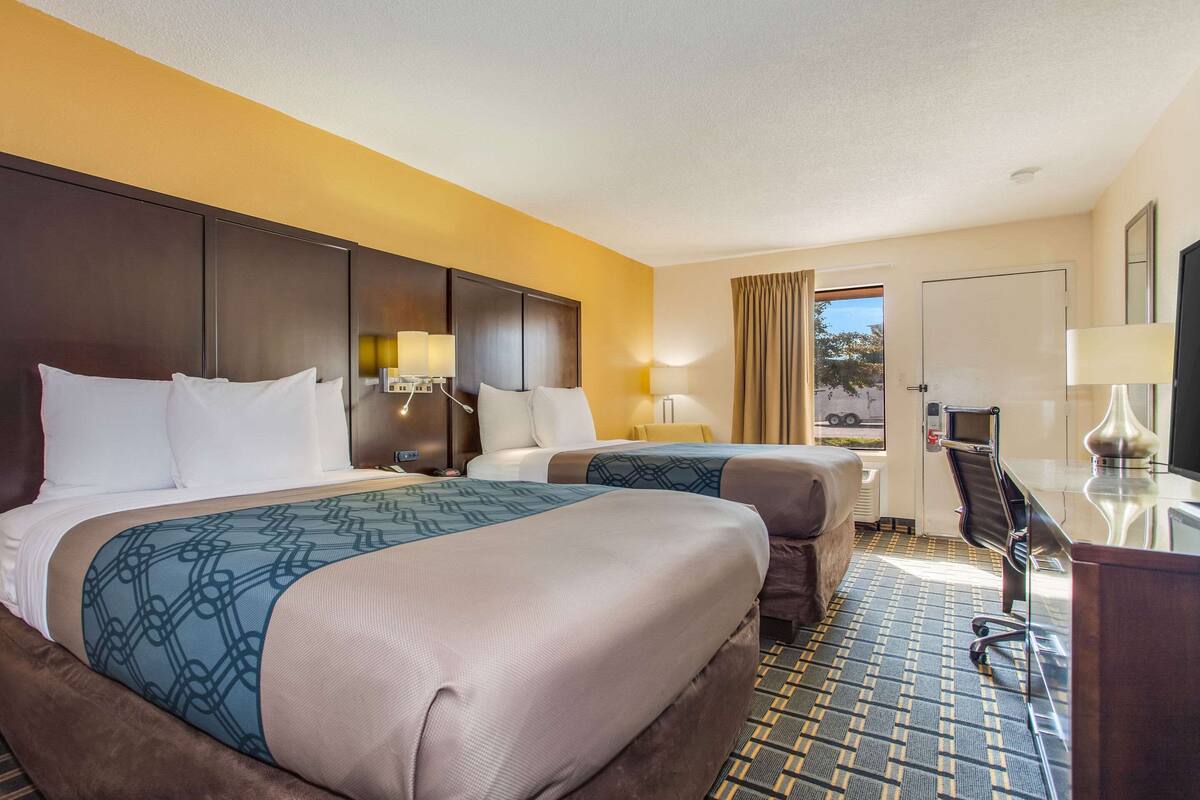 A hotel room with two beds, a desk, and a chair. There is a window with curtains, a door, and a lamp on the desk.