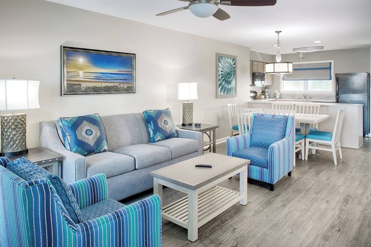A modern living room with a grey sofa, striped armchairs, a coffee table, and a dining area with white chairs.