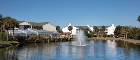 A serene lakeside view with a fountain, surrounded by white buildings and palm trees.