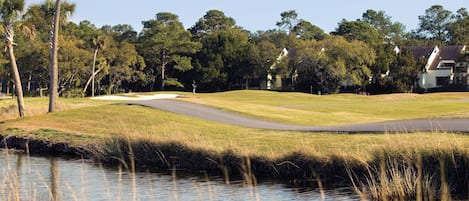 A golf course with a water body in the foreground, surrounded by trees and a building in the background.