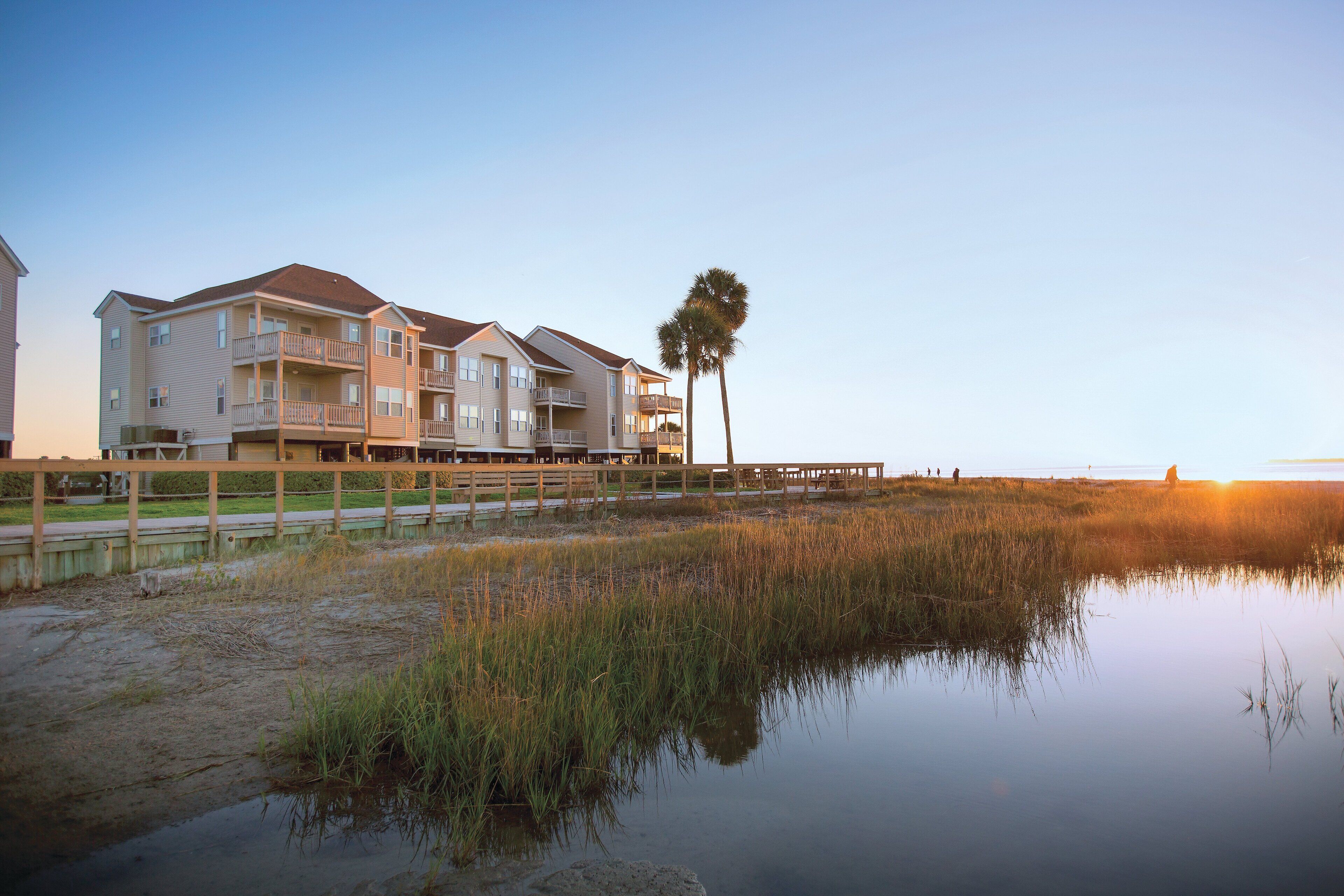 A beachfront apartment complex with multiple balconies, a wooden deck, and a view of the sunset over the water.