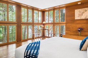 A bedroom with a large bed, wooden floor, and a view through large windows.