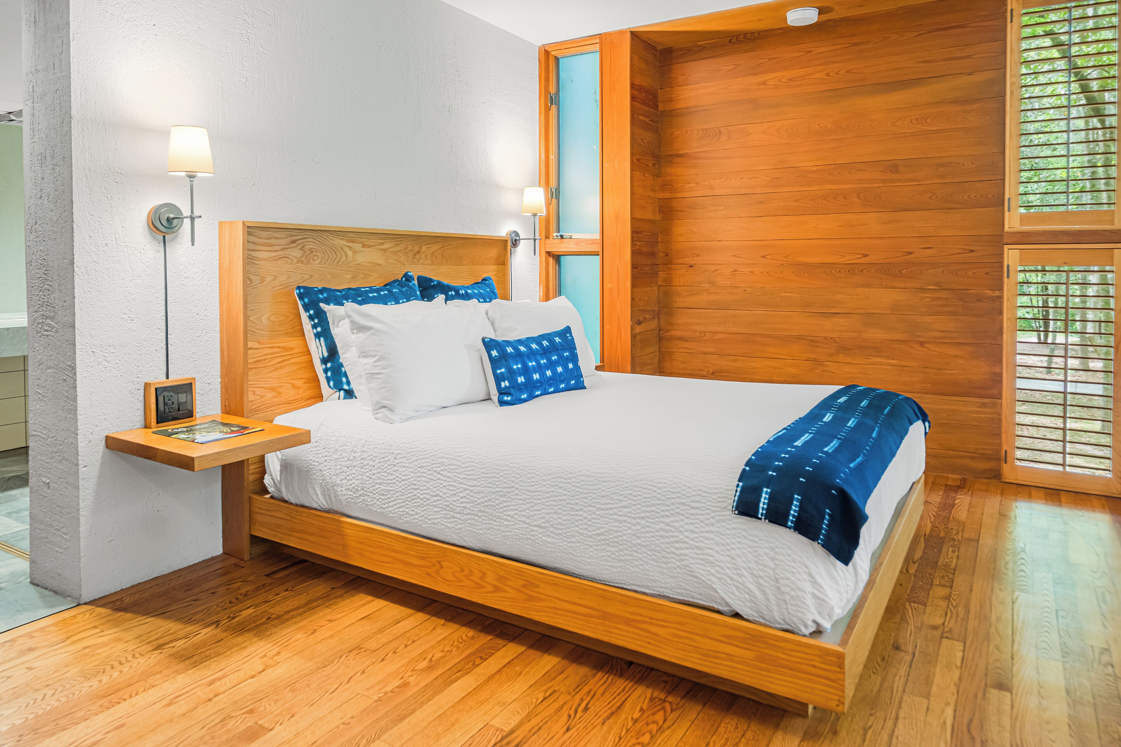 A wooden-framed bed with a white and blue bedspread, a nightstand with a lamp, and a window with wooden blinds.