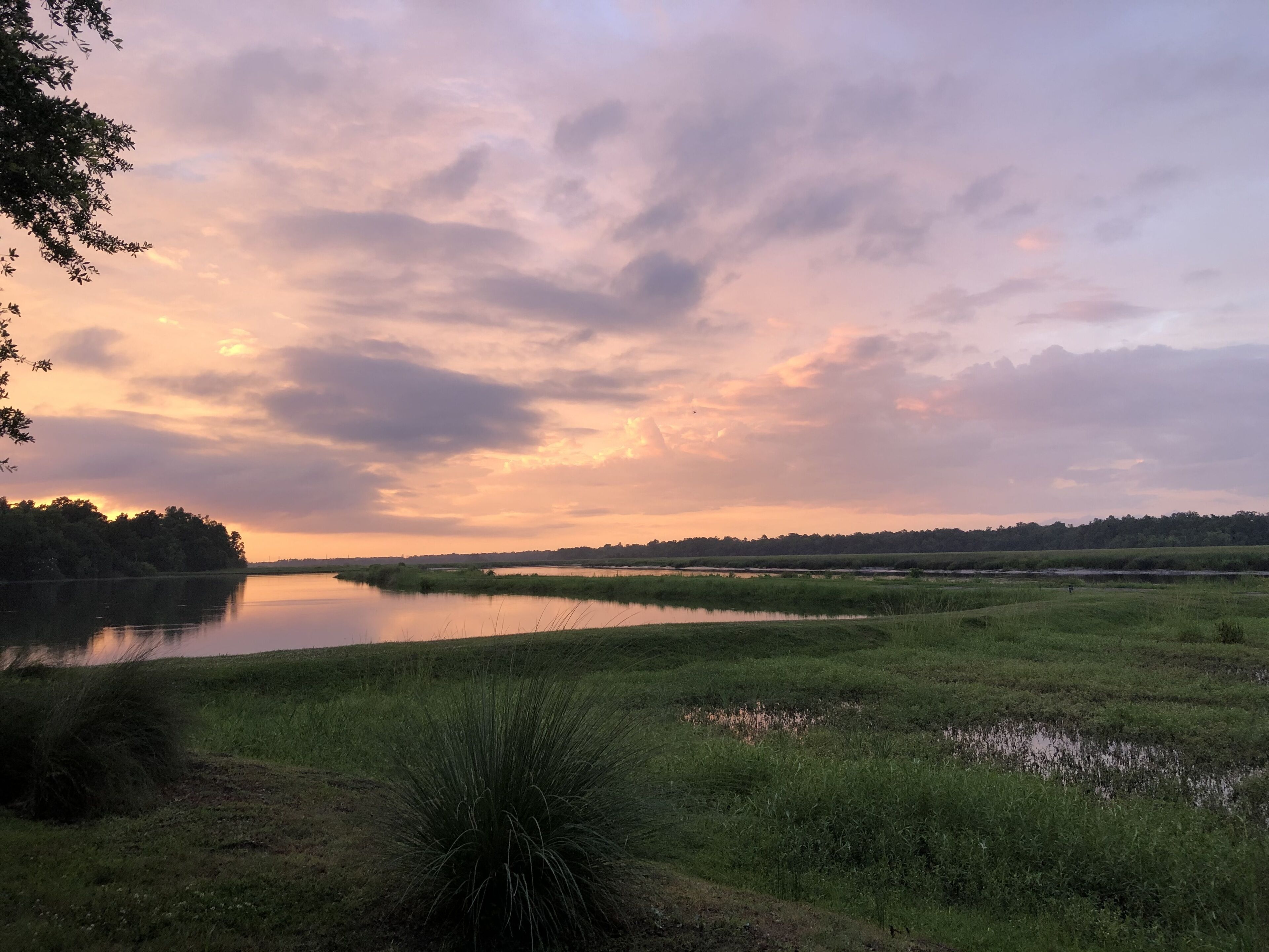 A serene landscape with a lake, grassy fields, and a distant tree line under a colorful sky.