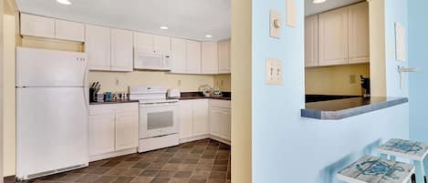 A kitchen with white cabinets, a refrigerator, a microwave, and a stove. There are two stools at a counter.