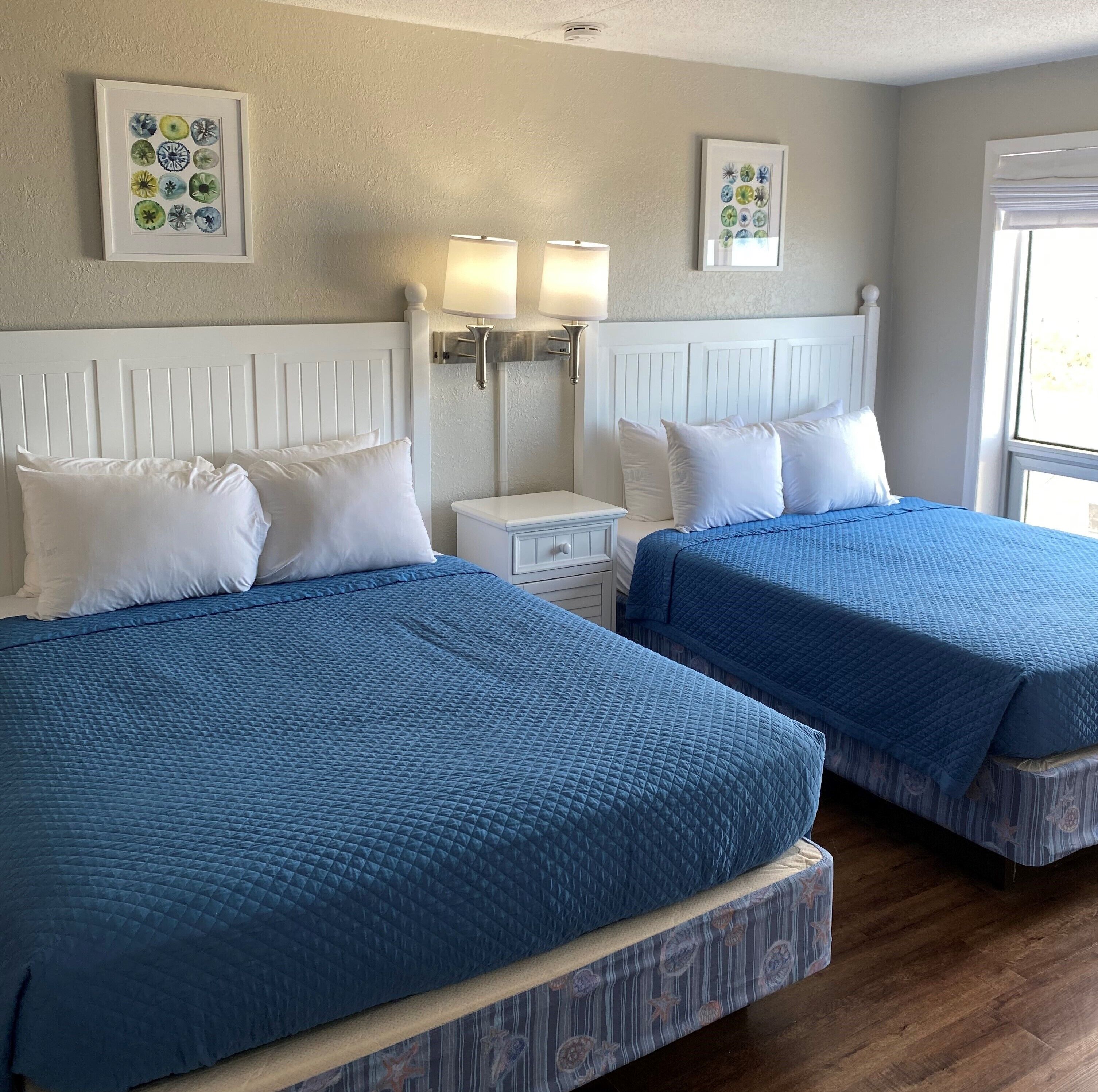 Une chambre d’hôtel avec deux lits, chacun doté de draps bleus et d’une tête de lit blanche. On y trouve deux tables de chevet avec des lampes, et des œuvres d’art sont accrochées aux murs.
