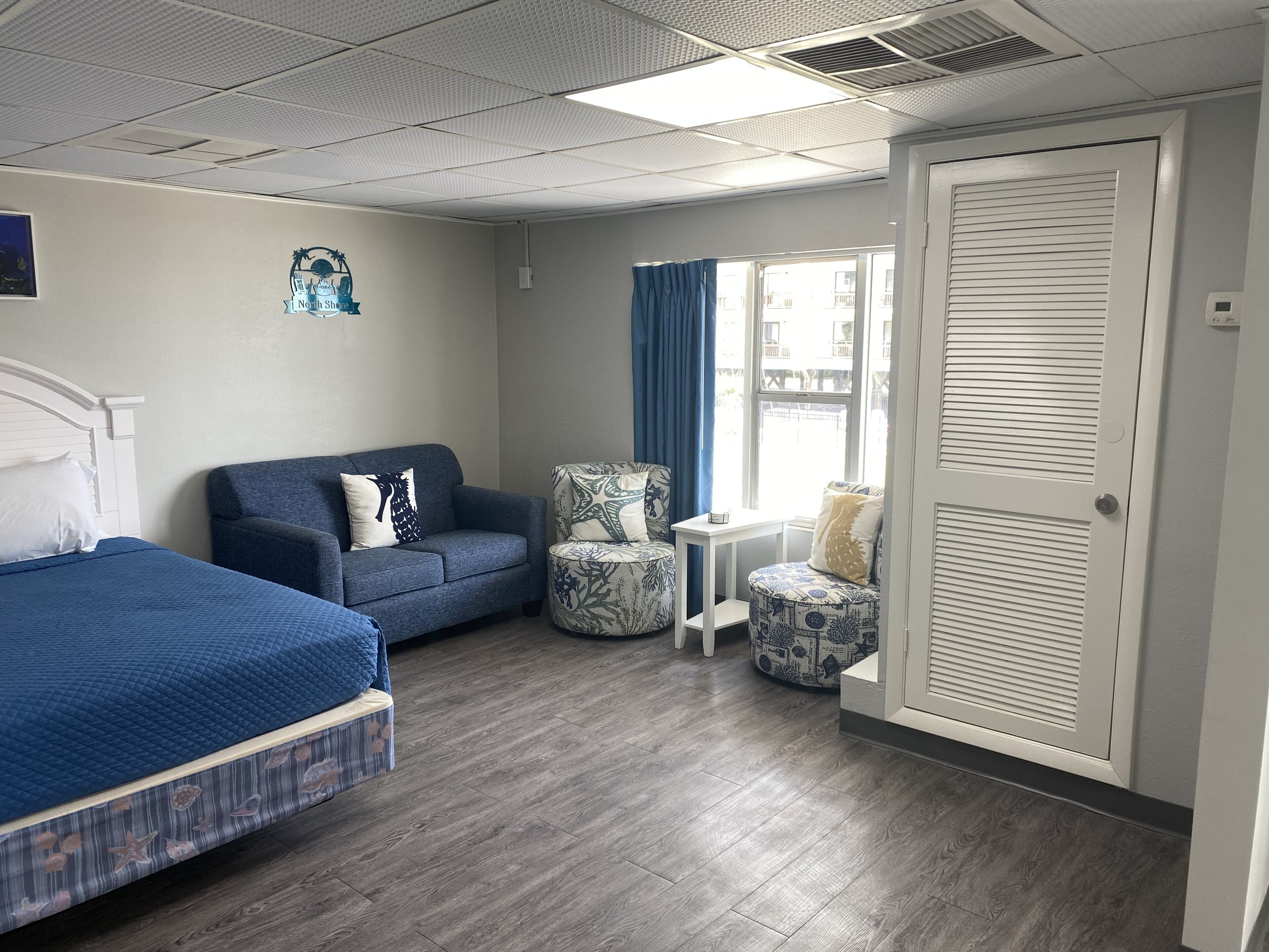Una habitación de hotel con cama, sofá, sillón, mesita y ventana con cortinas.
