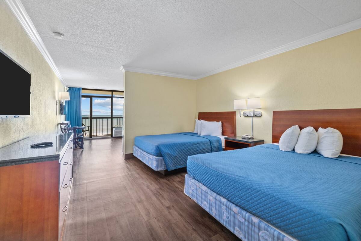 Ein Hotelzimmer mit zwei Betten, einem Schreibtisch, einem Stuhl, einem Fernseher und einem Balkon mit Blick.