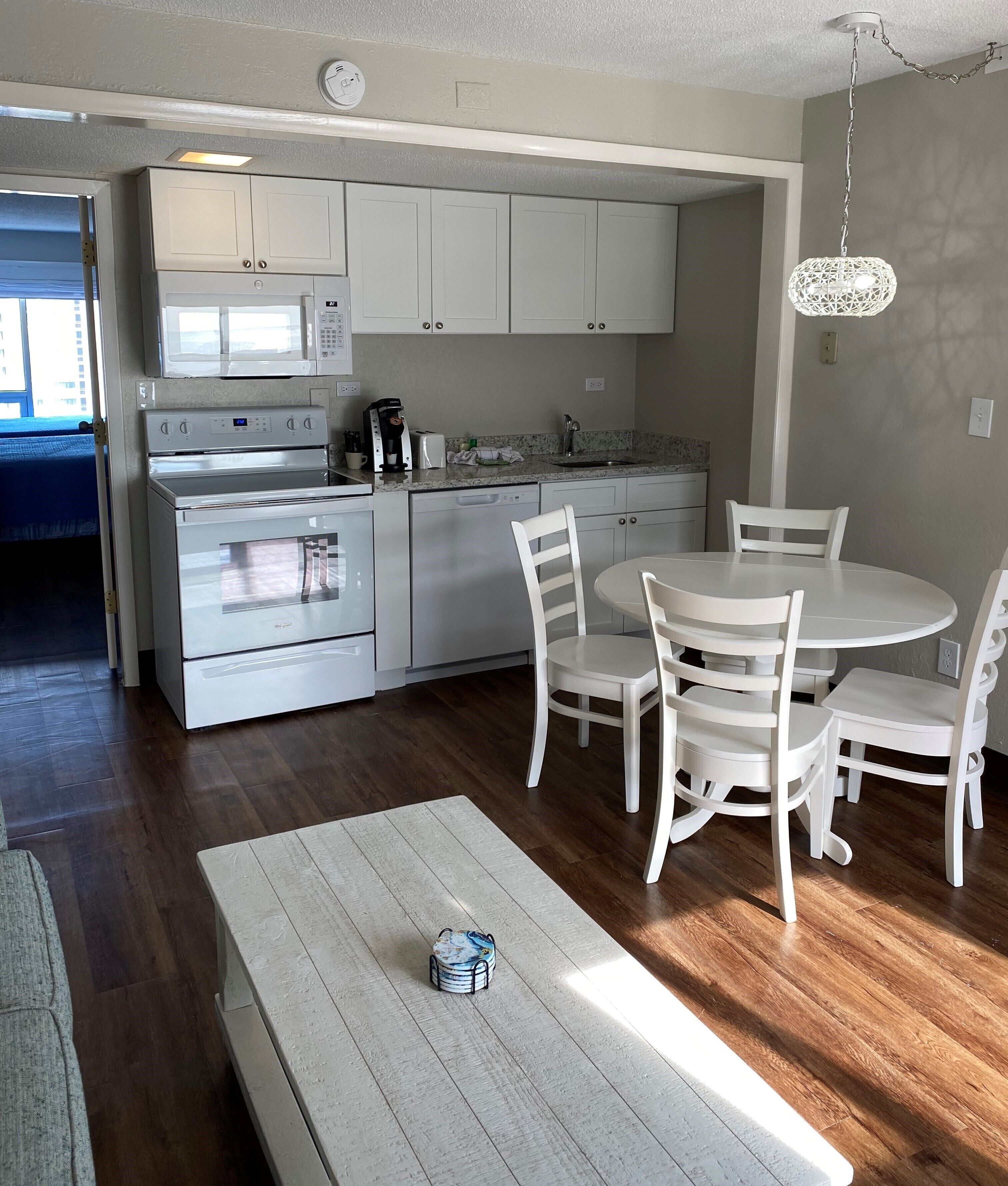A modern kitchen with white cabinets, a built-in microwave, a stove, and a dishwasher. There is a dining table with white chairs and a hanging light fixture.