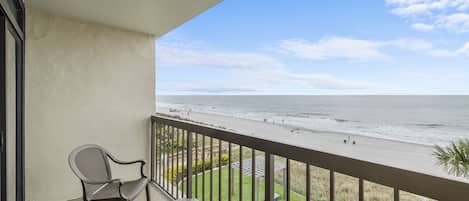 A balcony with a view of the beach, a chair, and a table.