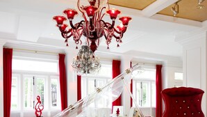 Un piano de cola en una habitación amplia con una araña, cortinas rojas y un sillón rojo.