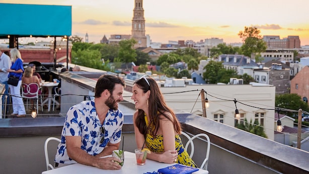 Una pareja cenando en un restaurante en la azotea, con el horizonte de la ciudad y la aguja de una iglesia al fondo.