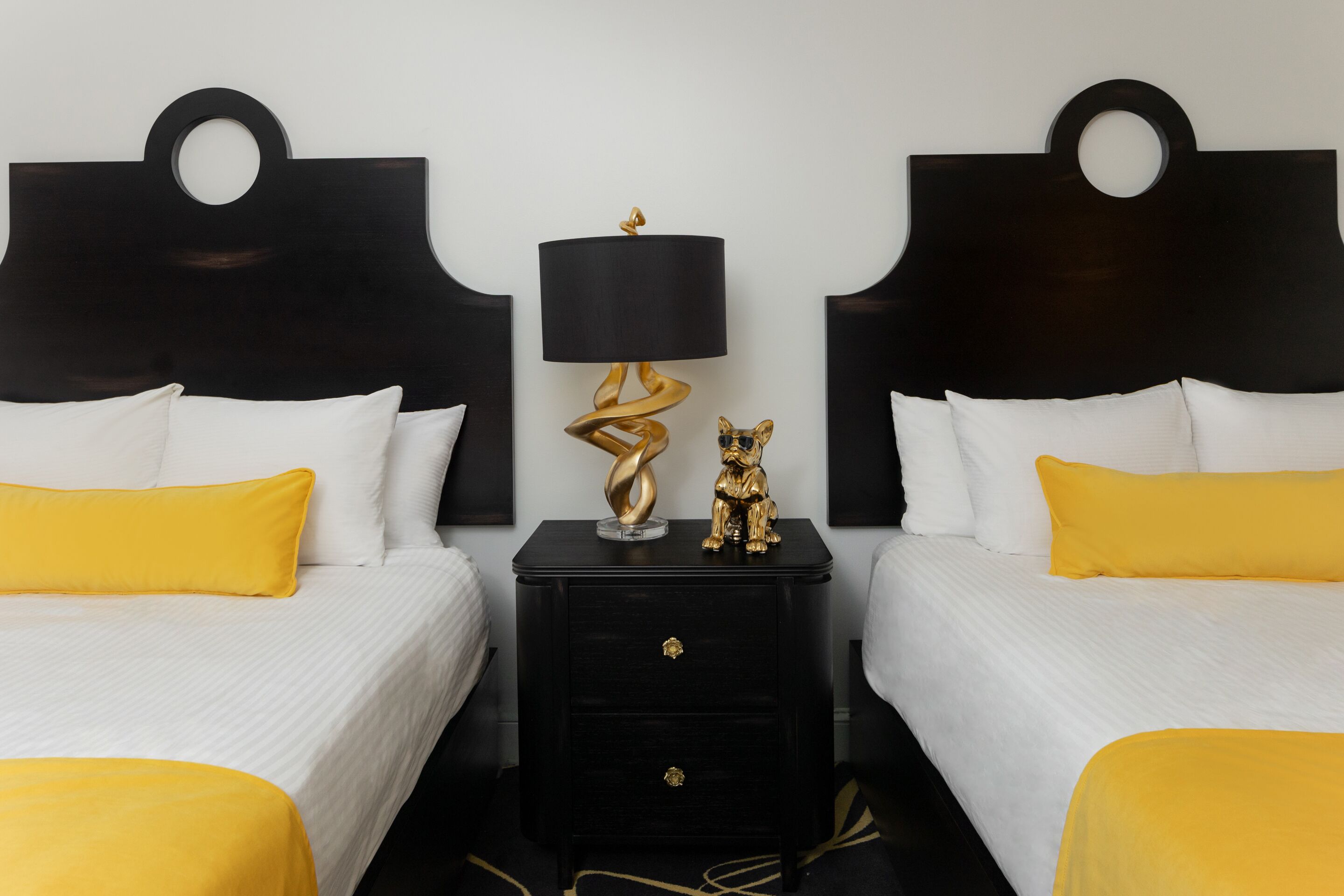 A hotel room with two beds, each with white bedding and yellow pillows. There is a black nightstand between the beds with a gold lamp and a small gold figurine.