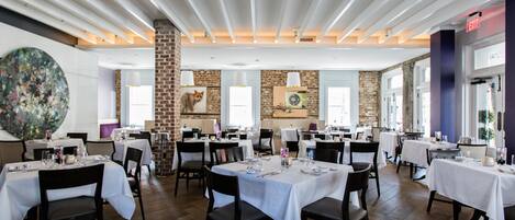 A spacious dining area with multiple tables set for dining, featuring a modern ceiling design and brick walls.
