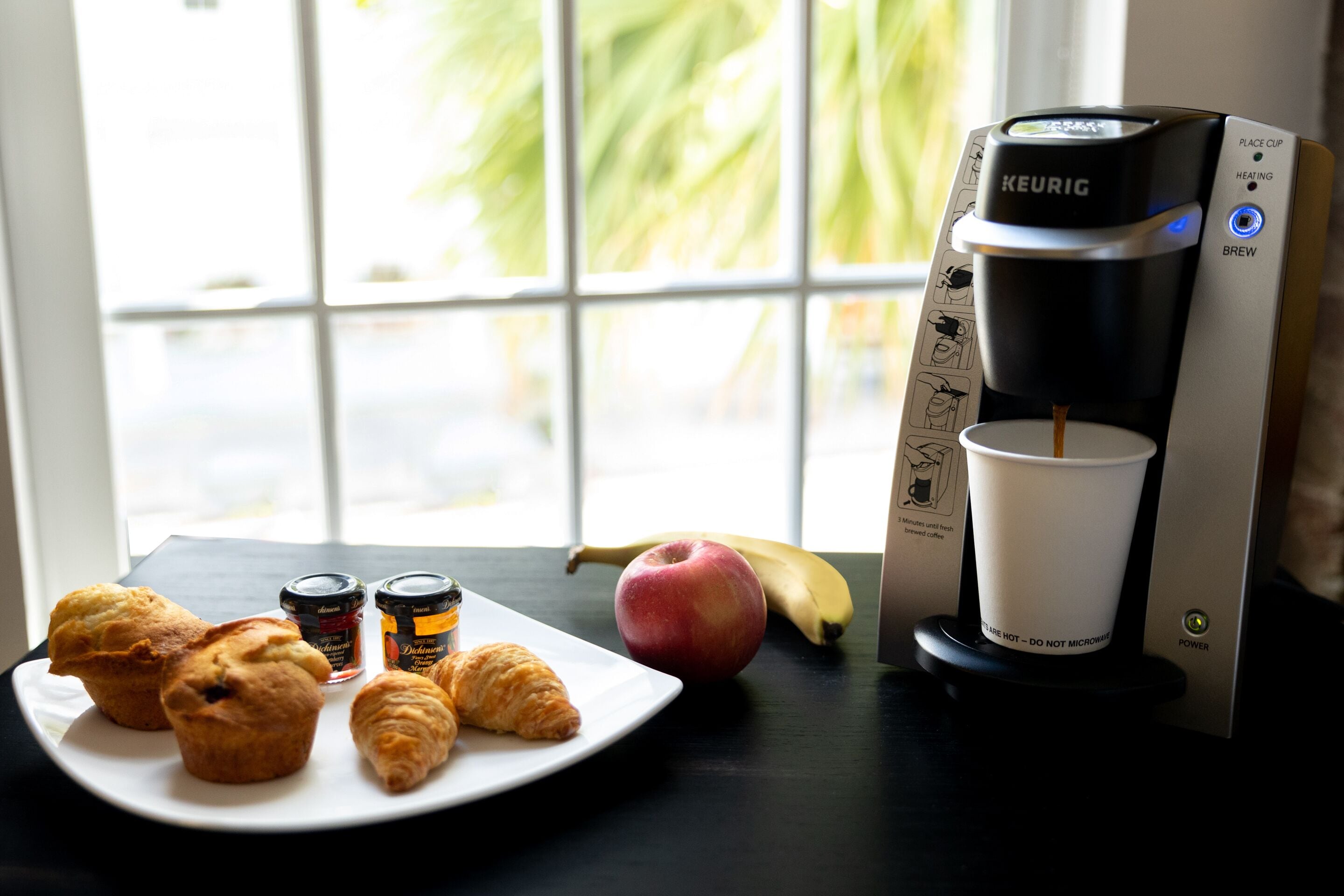 A Keurig coffee maker on a table with a plate of croissants, a banana, and an apple.