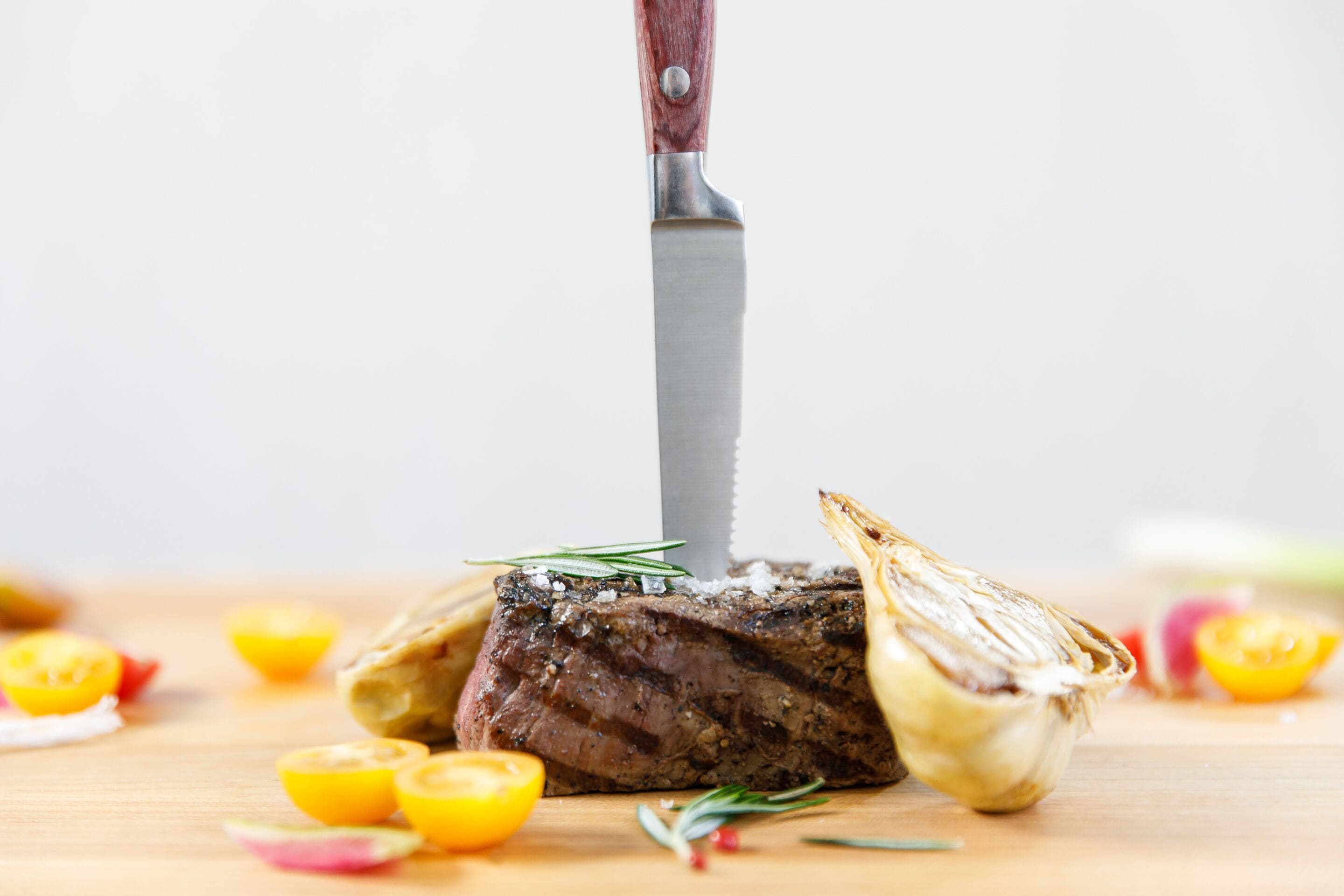 A steak with a knife inserted, surrounded by sliced tomatoes and a garlic bulb on a wooden surface.
