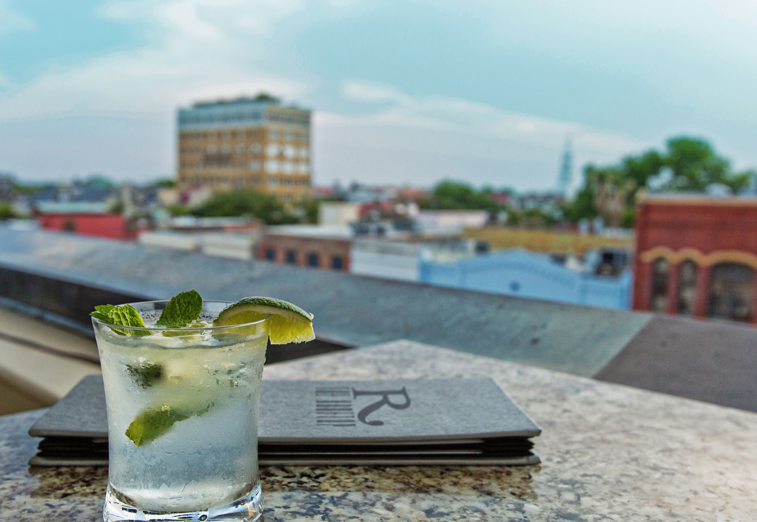 Un verre contenant une boisson claire, agrémentée de feuilles de menthe et d’une tranche de citron vert, est posé sur une table. En arrière-plan, on aperçoit une vue sur la ville.