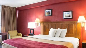 Una habitación de hotel con una cama, dos lámparas de noche, una silla, una mesita de noche con un reloj y dos cuadros en la pared.