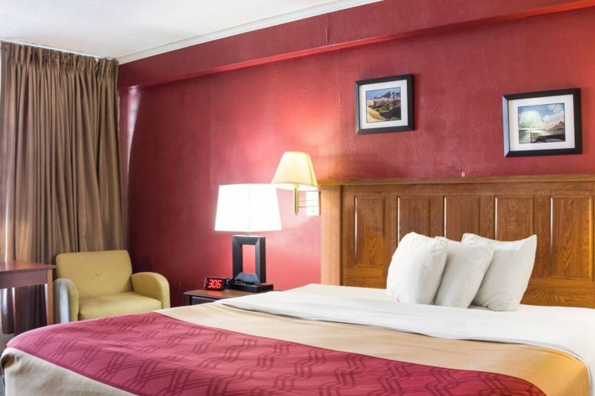Una habitación de hotel con una cama, dos lámparas de noche, una silla, una mesita de noche con un reloj y dos cuadros en la pared.