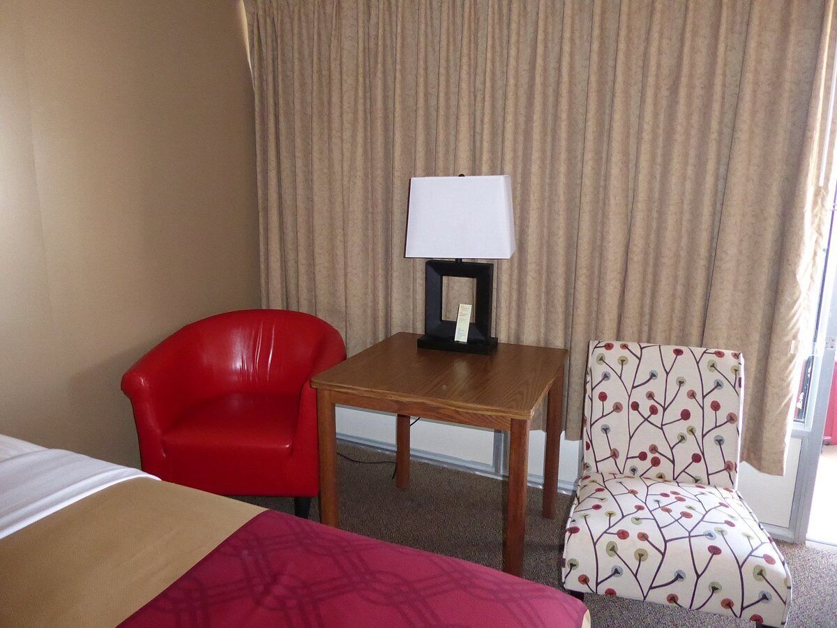 A hotel room with a bed, a red chair, a patterned armchair, and a small table with a lamp.