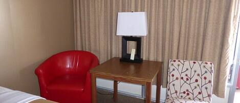 A hotel room with a bed, a red chair, a patterned armchair, and a small table with a lamp.