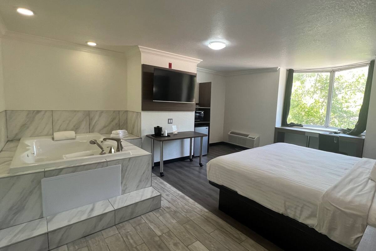 Une chambre d’hôtel moderne dotée d’un grand lit, d’une salle de bain séparée avec baignoire, d’une petite kitchenette et d’une fenêtre donnant sur la verdure.