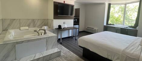 Habitación de hotel moderna con una cama grande, baño separado con bañera, pequeña kitchenette y ventana con vistas a árboles.