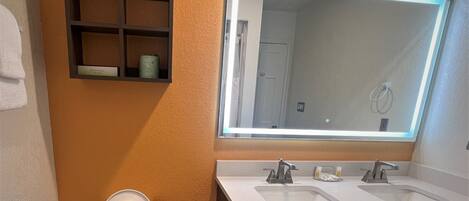 Un baño con inodoro blanco, dos lavamanos, un espejo grande y un estante con artículos de aseo.